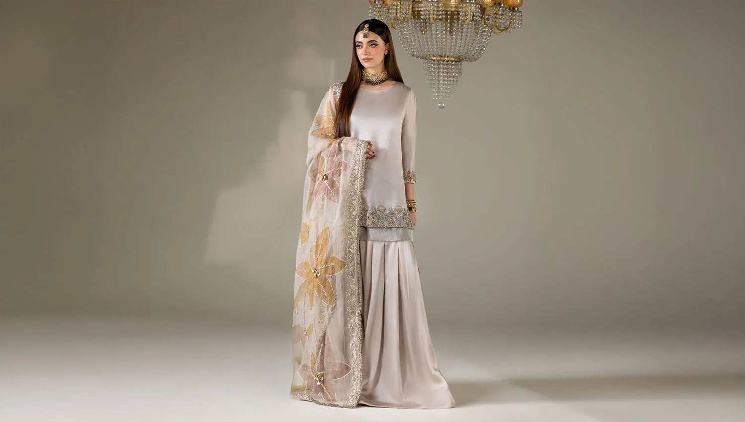 A woman models a traditional South Asian outfit consisting of a light beige long-sleeve kurta with silver embroidery, a matching pleated skirt, and a sheer beige dupatta with floral prints and silver borders, set against a neutral studio background with a hanging chandelier.