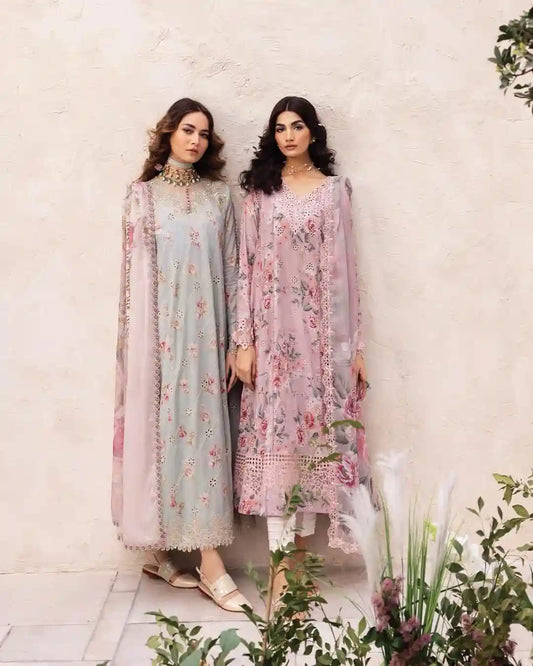 Two women modeling traditional South Asian salwar kameez suits in a garden setting against a textured beige wall. The woman on the left wears a light blue-green kameez with pink floral embroidery, and the woman on the right wears a dusty pink kameez with a pink floral print.